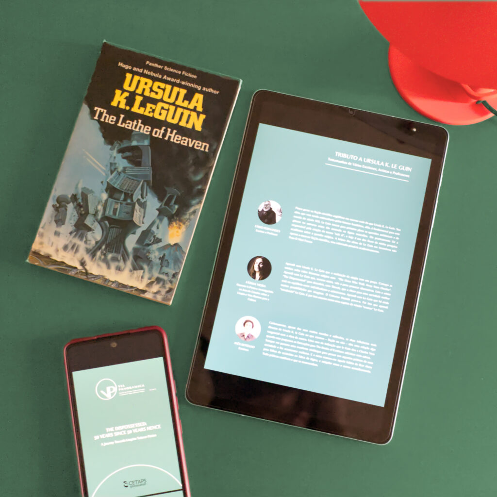 A phone, a tablet, and a book are arranged atop a deep, forest green background. The phone displays the teal-green front cover of the 2024 summer issue of academic journal Via Panoramica, while the tablet shows an excerpt with testimonials about Ursula K. Le Guin’s influence. This summer issue commemorates the 50th anniversary of Le Guin’s novel “The Dispossessed”, but the book in the image is Le Guin’s “The Lathe of Heaven”. In the image’s upper right corner, a modern orange-red table lamp contrasts with the greens.